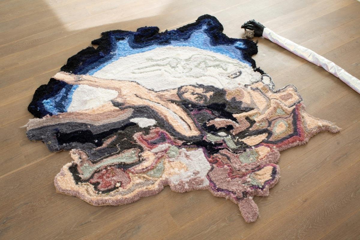 A wool rug on a hardwood floor, with a variety of colours including navy, blue, white and earthy colours. An abstract figure of a person in it can be seen in the rug.