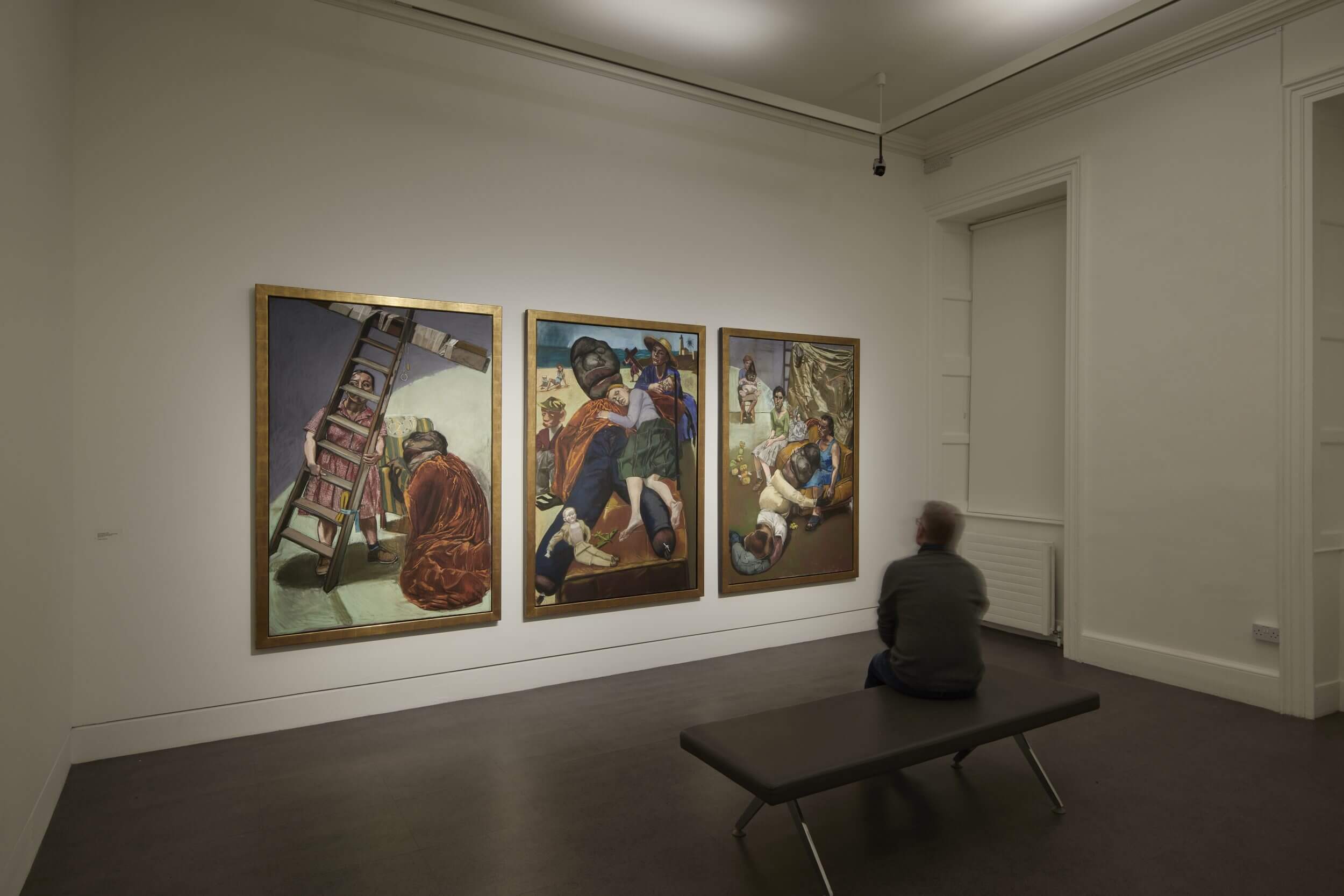 Interior installation photograph inside a gallery room with three large oil paintings on the wall. A person sits on a bench looking at the paintings.