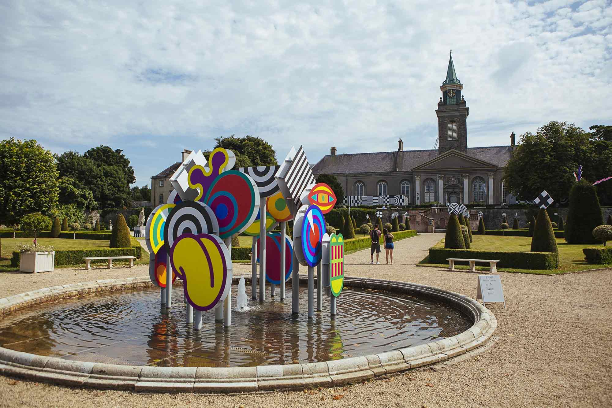 Exterior installation photograph of a fountain in a formal garden, an installation of colourful shapes in the centre of the fountain.