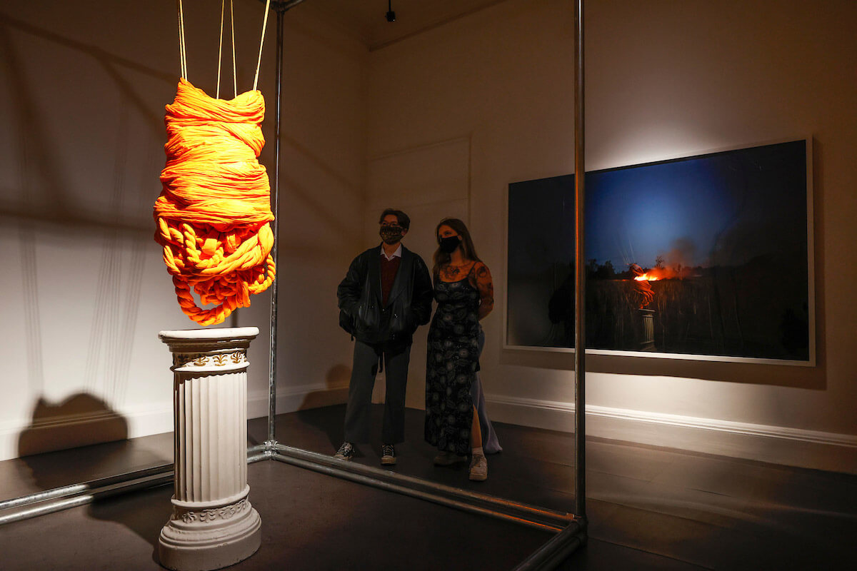 Interior gallery room photograph of two people looking at an installation with a large, framed picture on the wall behind them. The installation of orange cloth hung from the ceiling over a column plinth that is spotlighted