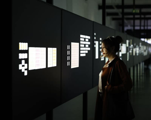 Interior installation photograph inside a long dark corridor, with display panels showing illuminated stamps. A woman stands is looking at the display.