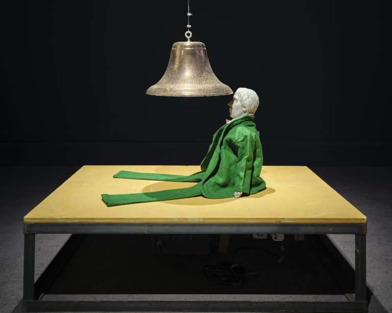 Installation of a cast iron bell suspended from ceiling, above a figurine in a green felt suit seated on a wooden base