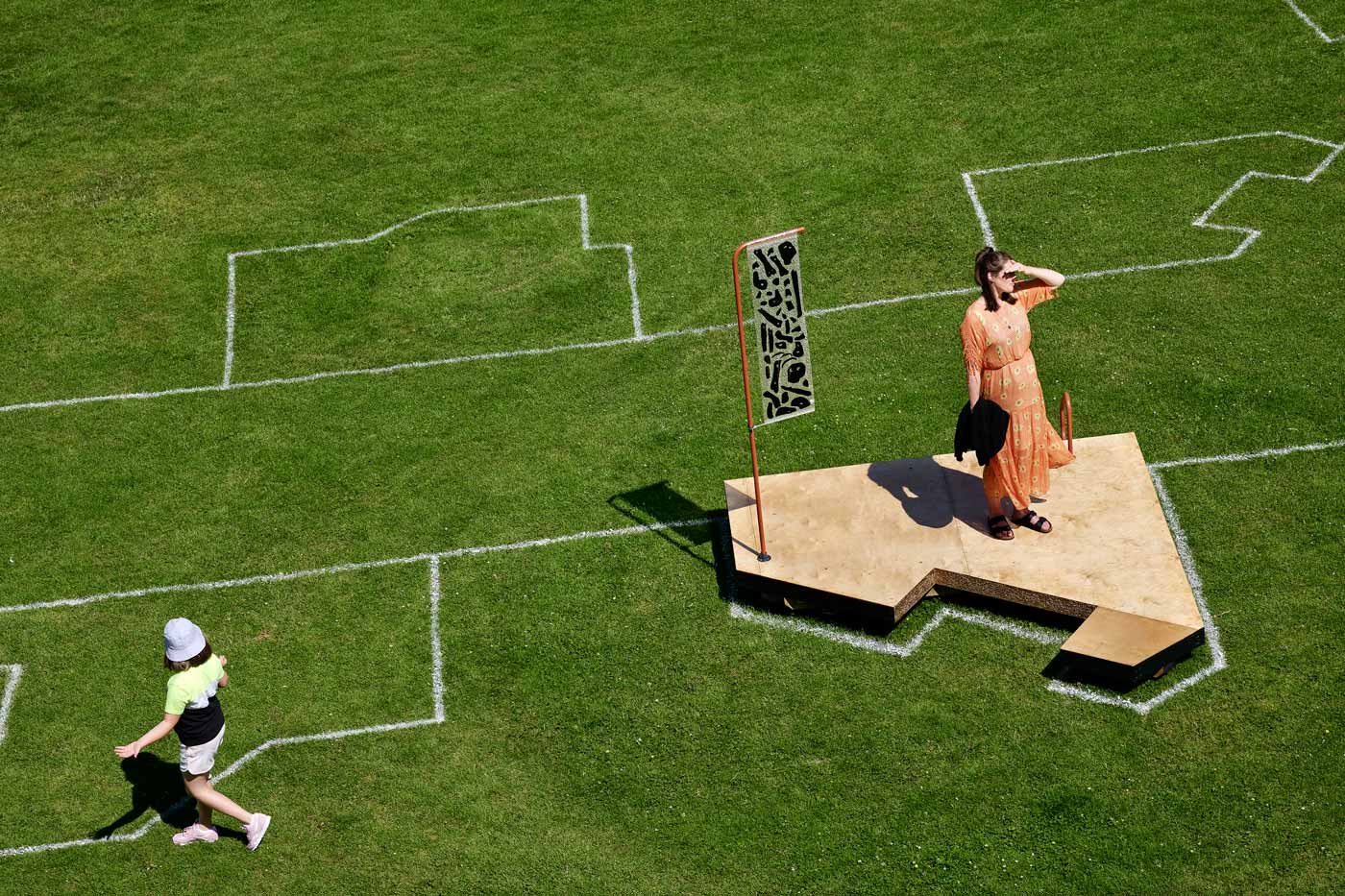Exterior installation photograph of a green lawn with white geometric lines painted on the grass. A woman stands on a wooden installation seating structure