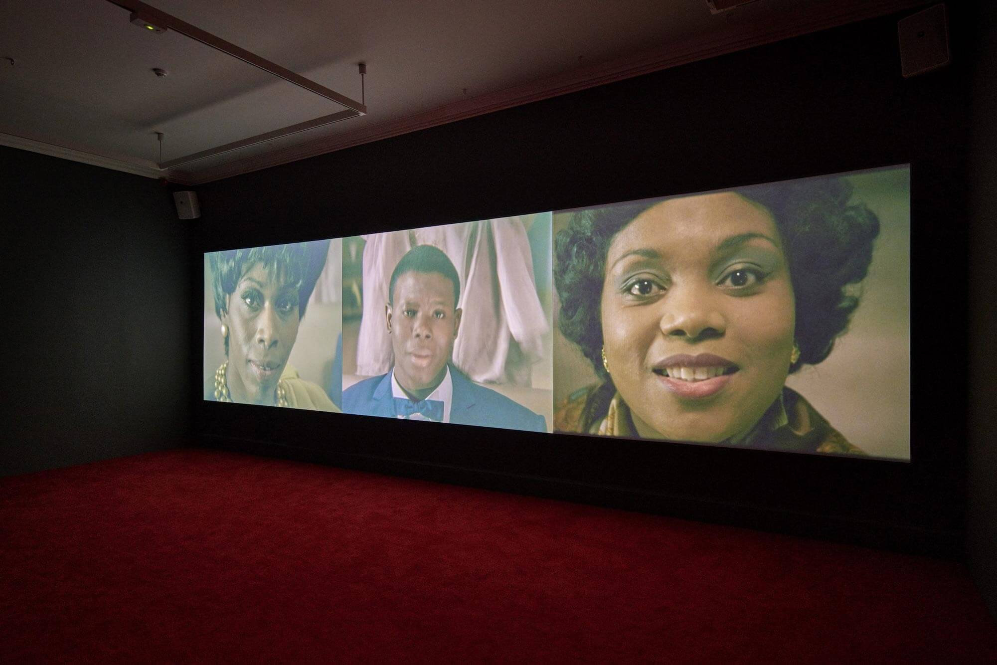 Interior photograph of a large screen showing a video installation. The screen is split into three sections, featuring a young black man and two black women