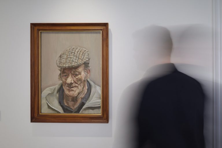Interior gallery room photograph of a framed painting of an older man in a peaked cap. A person stand in front of the painting, blurred as they are moving