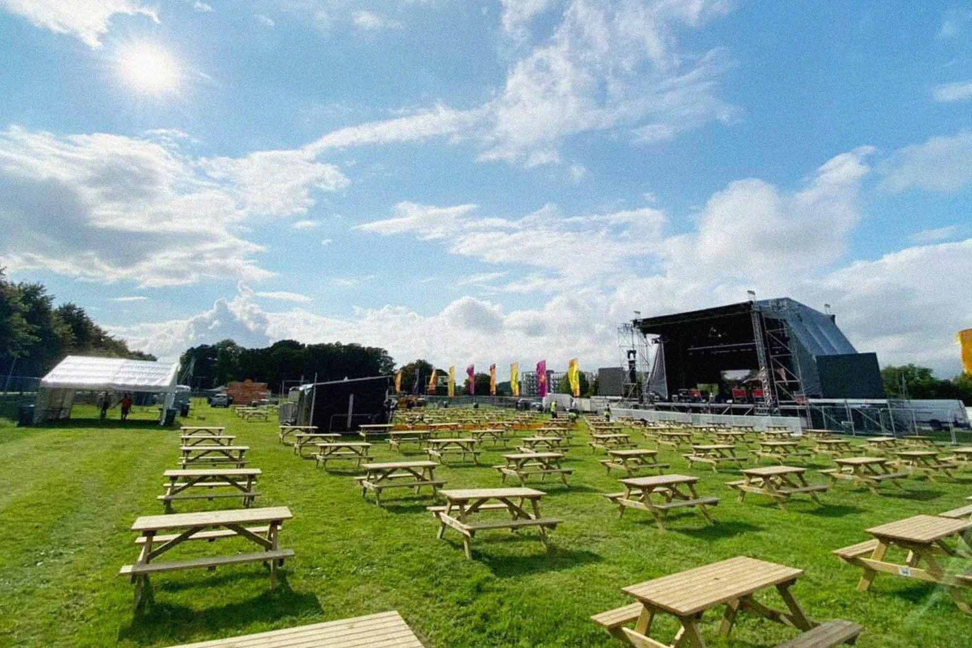 Photograph of a concert space set up in the meadows on a sunny day. There are several rows of picnic tables in front of a large sound stage.