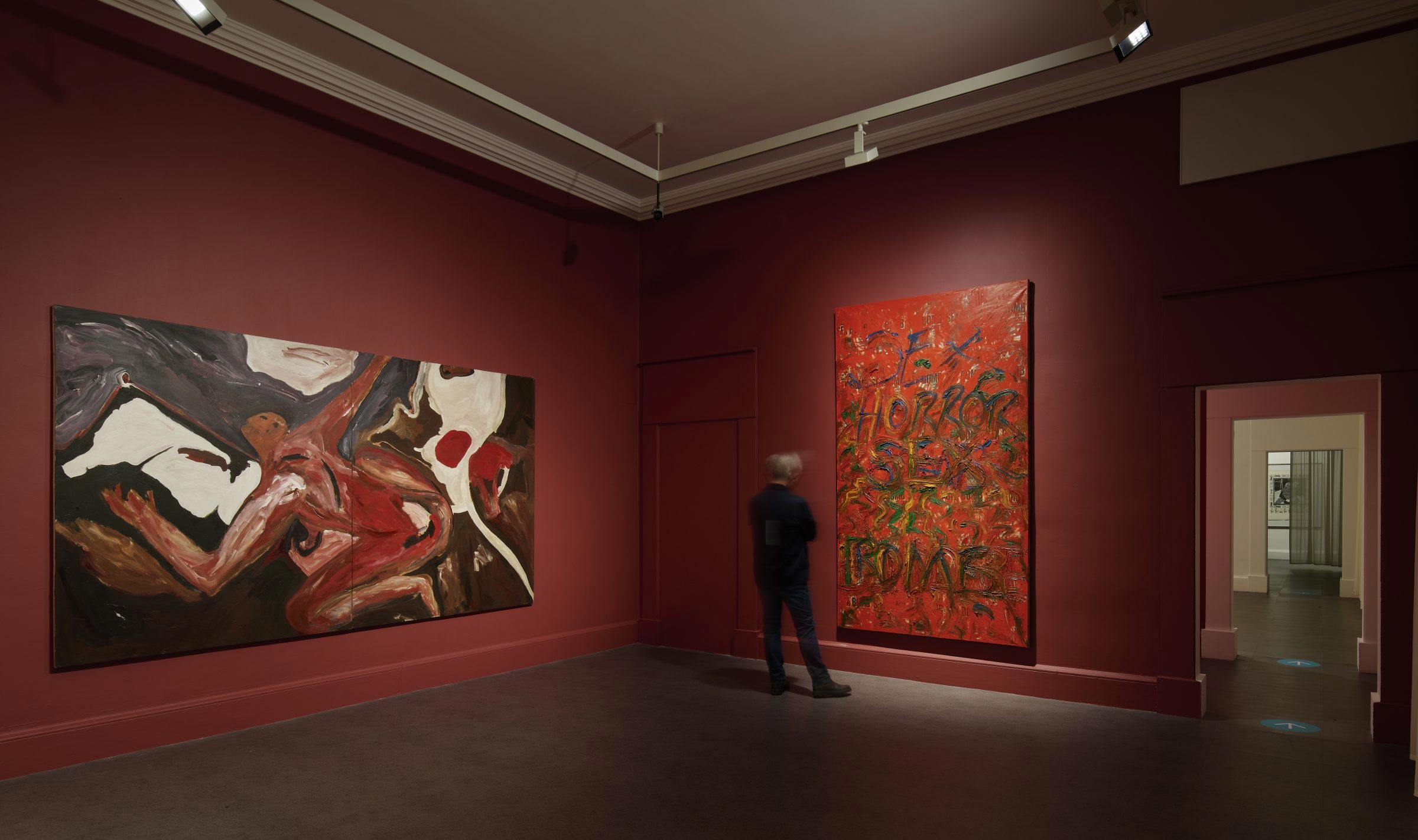 Interior photograph inside a gallery room with two large paintings. A blurred figure is looking at the works.