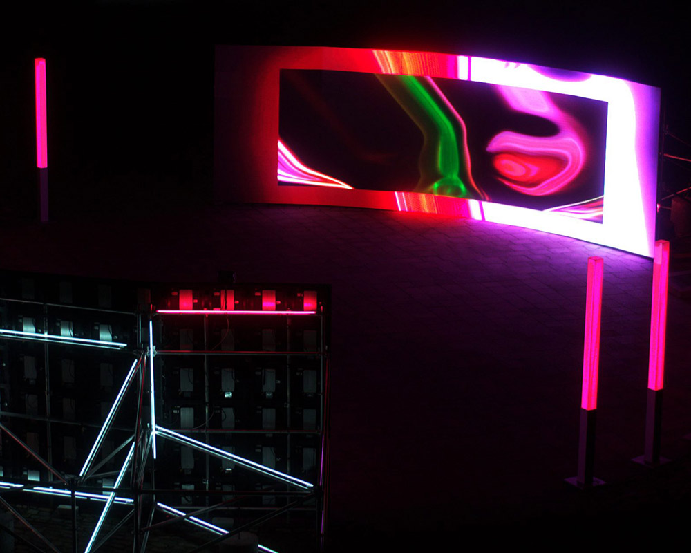 Night exterior installation photograph of a large screen displaying abstract neon colours. There are pink neon light columns on either side of the screen. In the foreground is the back of a second screen and it's supporting structure is illuminated.