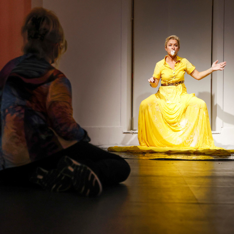 Interior installation photograph of a woman in a yellow dress, in the foreground of the photo is a figure seated on the floor looking at the performance.