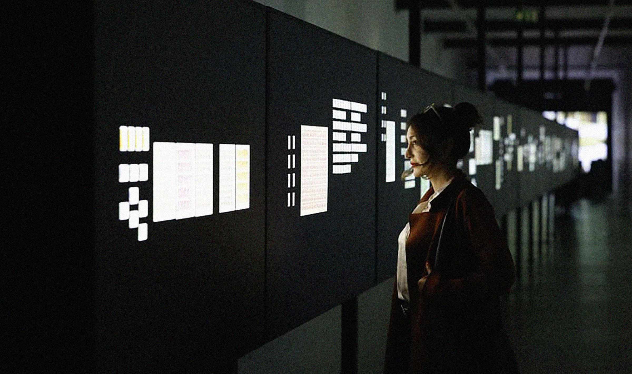 Interior installation photograph inside a long dark corridor, with display panels showing illuminated stamps. A woman stands is looking at the display.